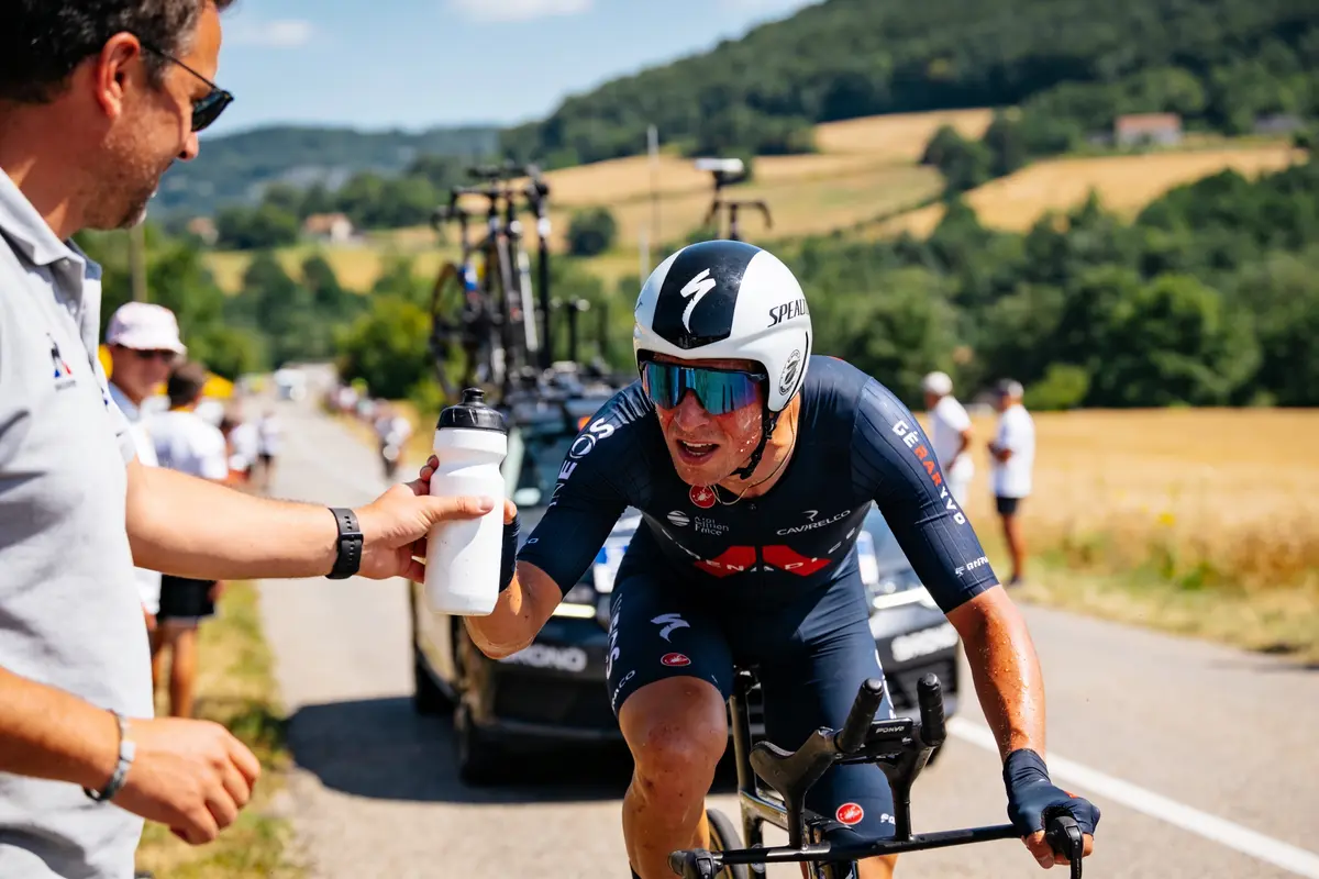 Cycliste recevant une bouteille d'eau lors d'une course en campagne, voiture d'assistance en arrière-plan.
