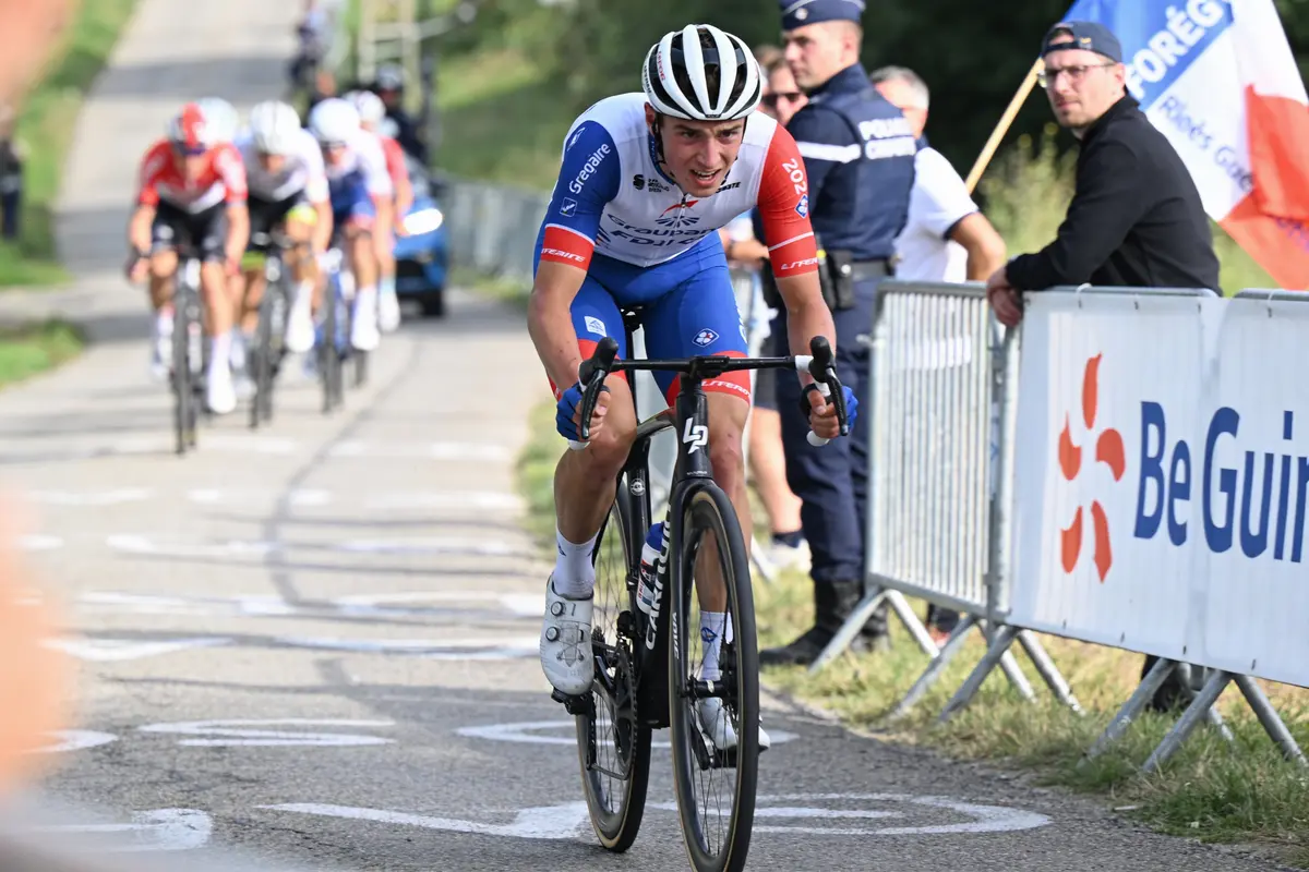 Cycliste en course sur route, suivi par d'autres, présence de spectateurs et policiers en arrière-plan.