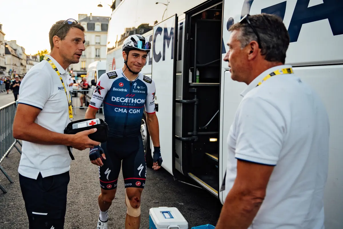 Cycliste en tenue sportive discutant avec deux hommes près d'un bus, équipement médical visible.
