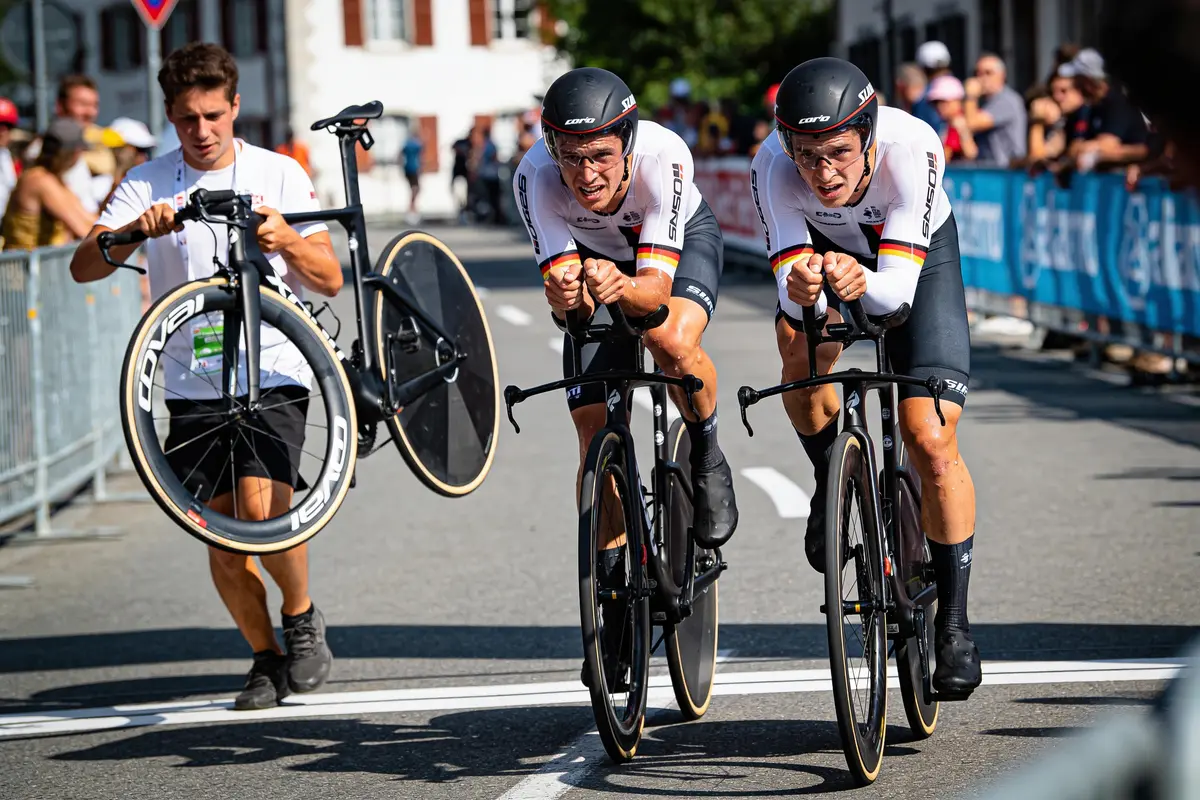 Deux cyclistes en course sur route, homme portant une roue de vélo en arrière-plan.