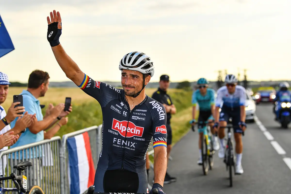 Cycliste saluant la foule, route de campagne, drapeau français, soutien des spectateurs, autres cyclistes en arrière-plan.
