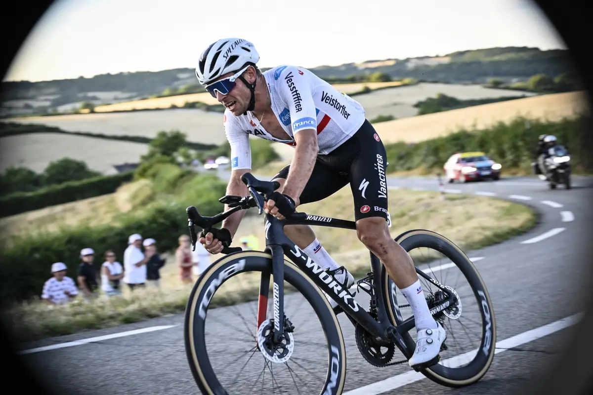 Cycliste en course sur une route de campagne sinueuse, avec spectateurs et voitures à l'arrière-plan.