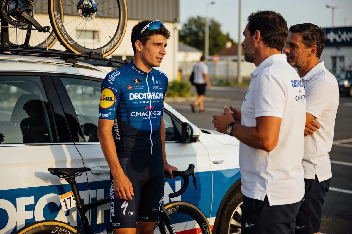 Cycliste en tenue de course discutant avec deux hommes devant une voiture d'assistance Decathlon.