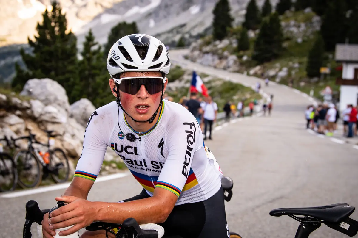 Cycliste avec casque et lunettes de soleil, route montagneuse, spectateurs en arrière-plan, drapeau français visible.