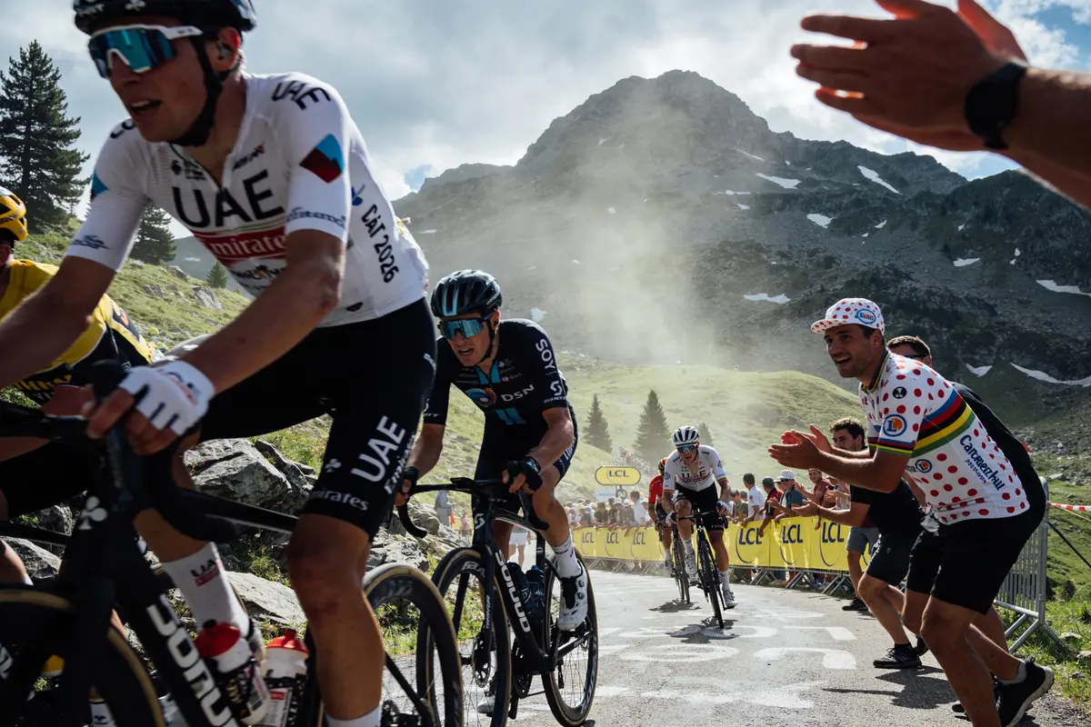 Cyclistes montant une route en montagne, entourés de spectateurs applaudissant, banderoles jaunes en arrière-plan.