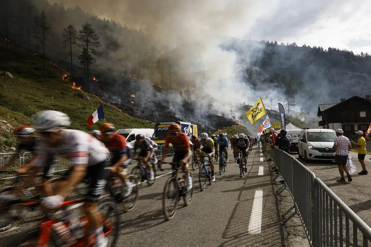 Cyclistes du Tour de France passent devant incendie de forêt sur une route sinueuse en montagne, drapeau français visible.