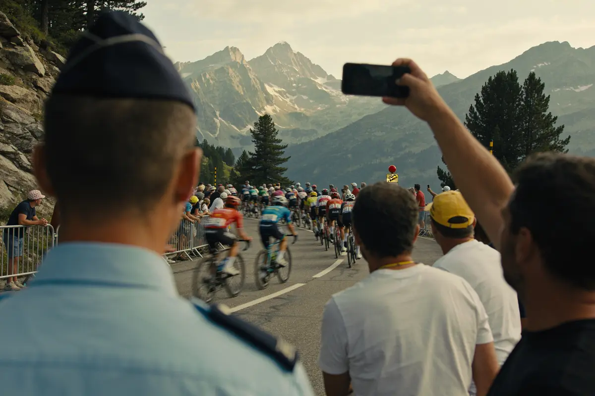 Course cycliste en montagne avec spectateurs, montagnes en arrière-plan sous un ciel partiellement nuageux.