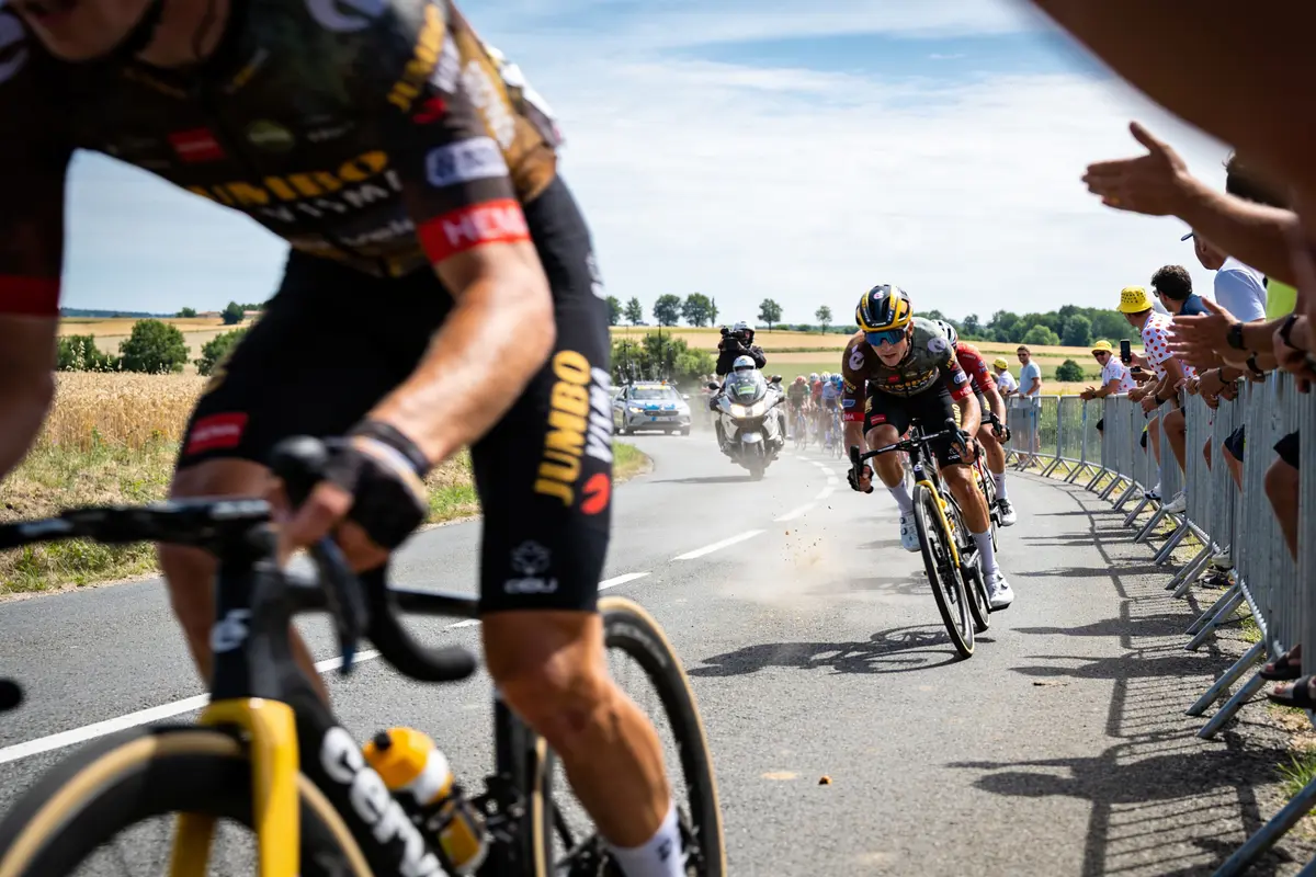 Cyclistes en course, encadrés par spectateurs, sur route de campagne ensoleillée.