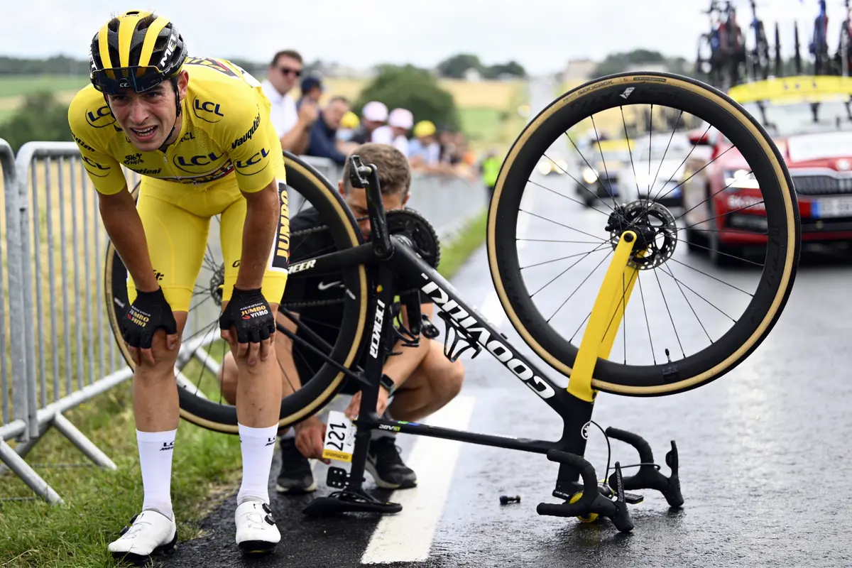Cycliste en tenue jaune debout près du vélo retourné, assistance en arrière-plan. Routes et spectateurs visibles.