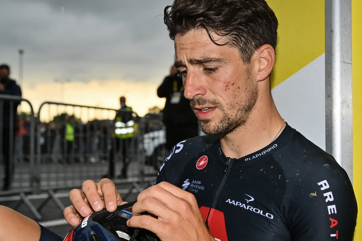 Cycliste essuyant du visage sale, casque à la main, fond flou de spectateurs et ciel nuageux.
