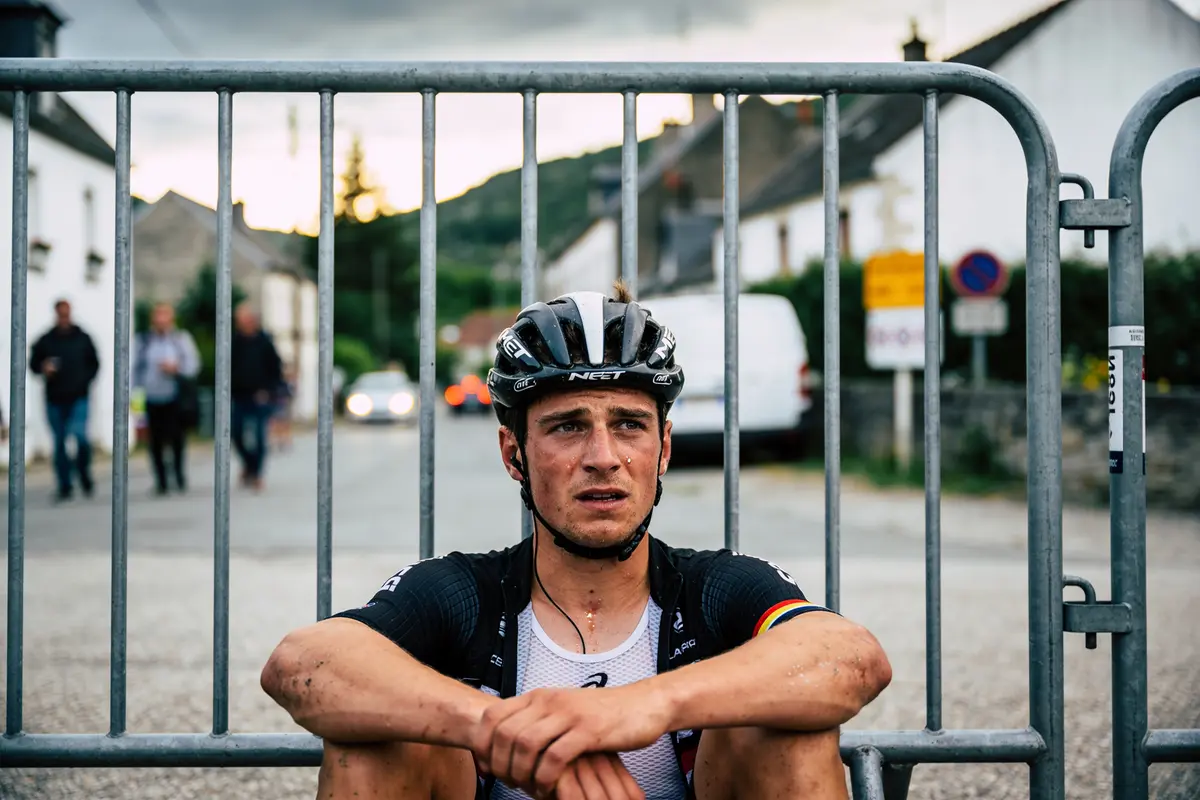 Cycliste assis derrière barrière, vêtu de noir, air fatigué, route de campagne en arrière-plan.