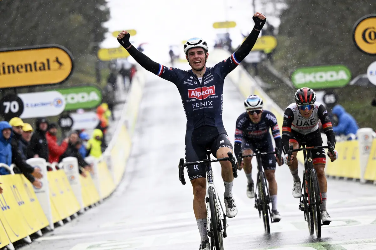 Cycliste levant les bras, franchissant la ligne d'arrivée sous la pluie, autres cyclistes derrière.