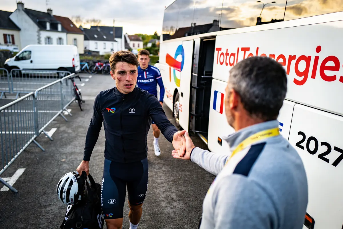 Cycliste saluant un homme devant un bus TotalEnergies, après une course, avec un coéquipier en arrière-plan.