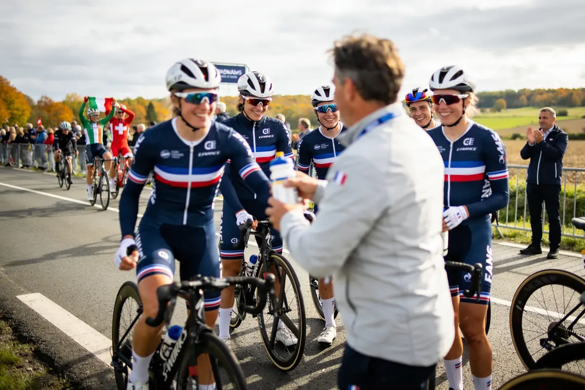 Coureuses cyclistes françaises souriantes, recevant des bouteilles en bord de route pendant une course, foule applaudissant.
