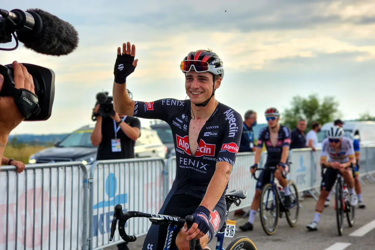 Cycliste souriant, saluant la caméra, en tenue Alpecin-Fenix, entouré de photographes et cyclistes, en extérieur.