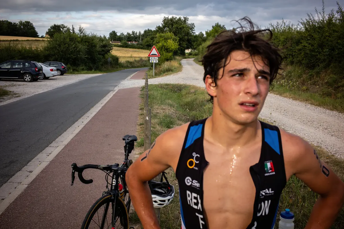Un athlète en maillot de triathlon, transpirant, se tient près de son vélo sur une route rurale.