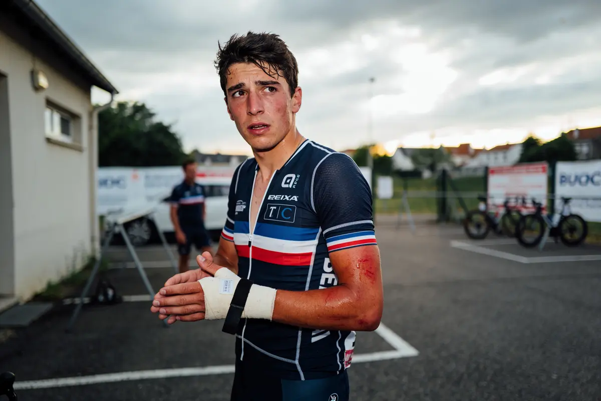 Cycliste blessé avec un bandage, montrant des marques rouges, devant un bâtiment par un ciel nuageux.