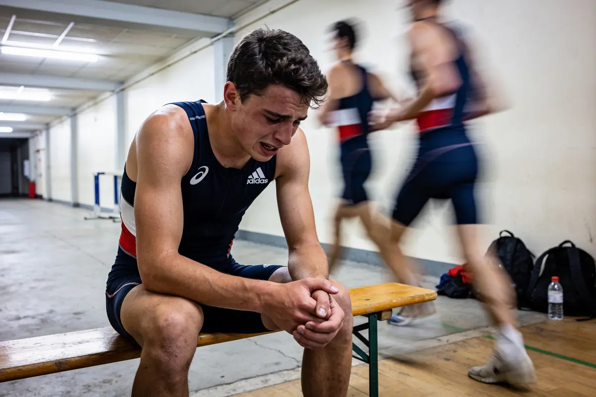 Athlète assis, en tenue de sport, semble épuisé, deux autres courent en arrière-plan flou.