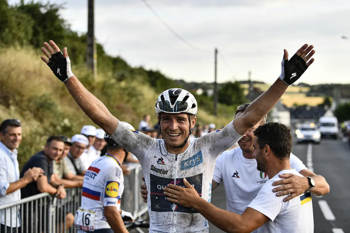 Cycliste souriant, bras levés, entouré de supporters après une course sur route, ciel dégagé, paysage rural.