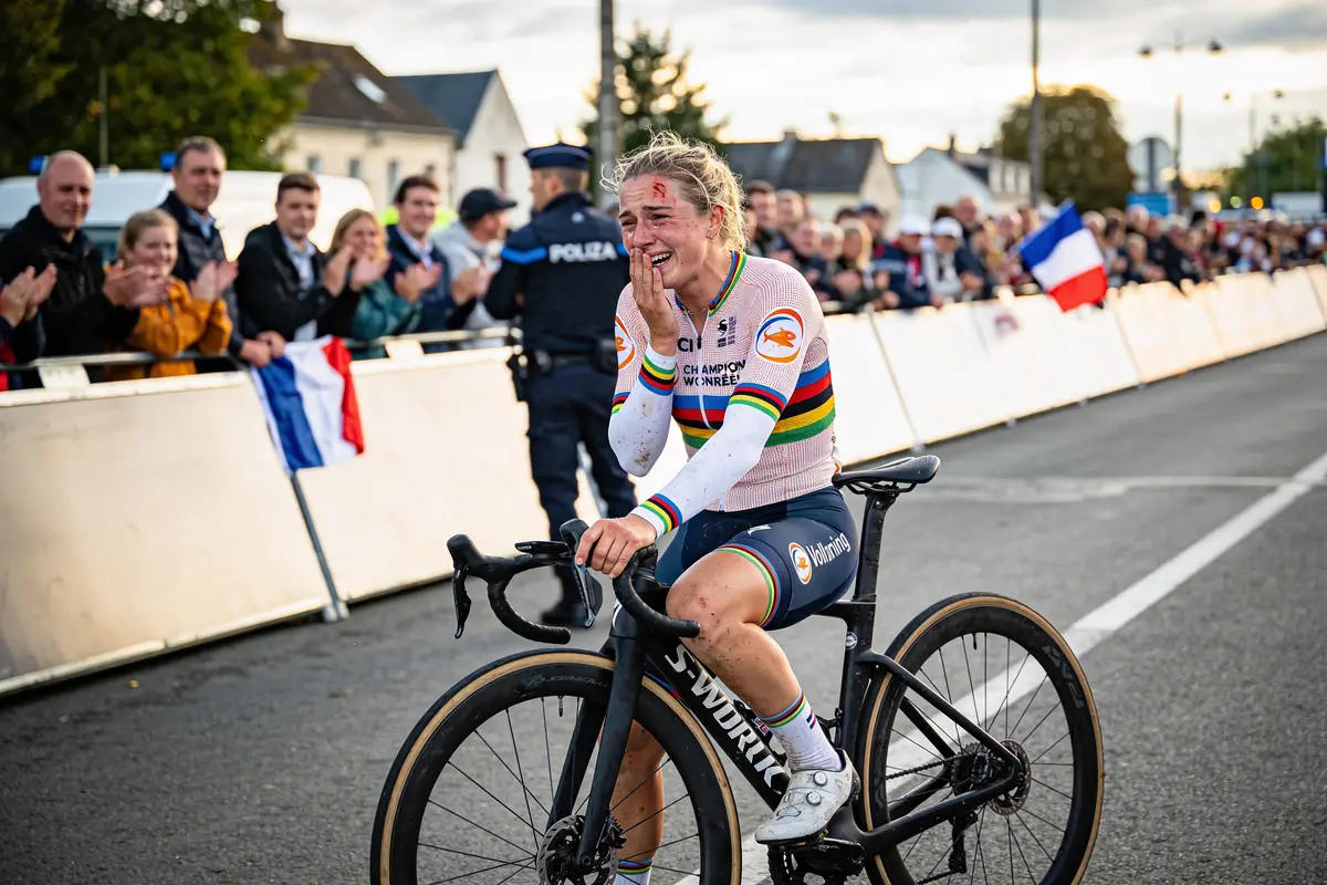 Cycliste souriante sur son vélo, applaudie par la foule sous un ciel nuageux. Drapeau français visible.